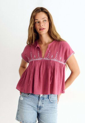 Femme aux longs cheveux portant un chemisier rose brodé ample et un short en jean clair, debout devant un fond uni et clair.