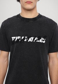 Czarny bawełniany t-shirt z okrągłym dekoltem, z zniszczonym białym napisem na wysokości klatki piersiowej. Krótkie rękawy i luźny krój.