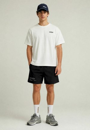 Jeune homme portant un t-shirt blanc avec le logo Cuera, un short noir avec des logos, des chaussettes blanches Cuera, des baskets grises New Balance et une casquette noire Cuera.