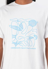 Tricou alb cu un design abstract de linii albastre. Textul de sub design spune "LIBERTATE DE LA HAOS." Guler rotund și mâneci scurte.