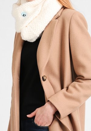 Écharpe tubulaire en grosse maille crème avec motif texturé, associée à un manteau beige à fermeture unique par bouton. Haut noir visible en dessous.