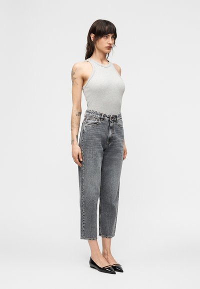 Γυναίκα με σκούρα μαλλιά που φοράει αμάνικη γκρι μπλούζα, ψηλόμεσα γκρι τζιν, μαύρα μυτερά παπούτσια, στέκεται μπροστά σε απλό λευκό φόντο.