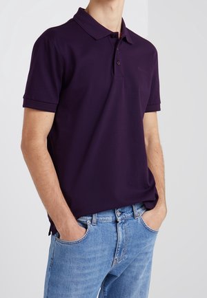 Hombre con una camisa polo de manga corta morada oscura, por fuera, sobre unos jeans azul claro, con las manos en los bolsillos, contra un fondo claro.