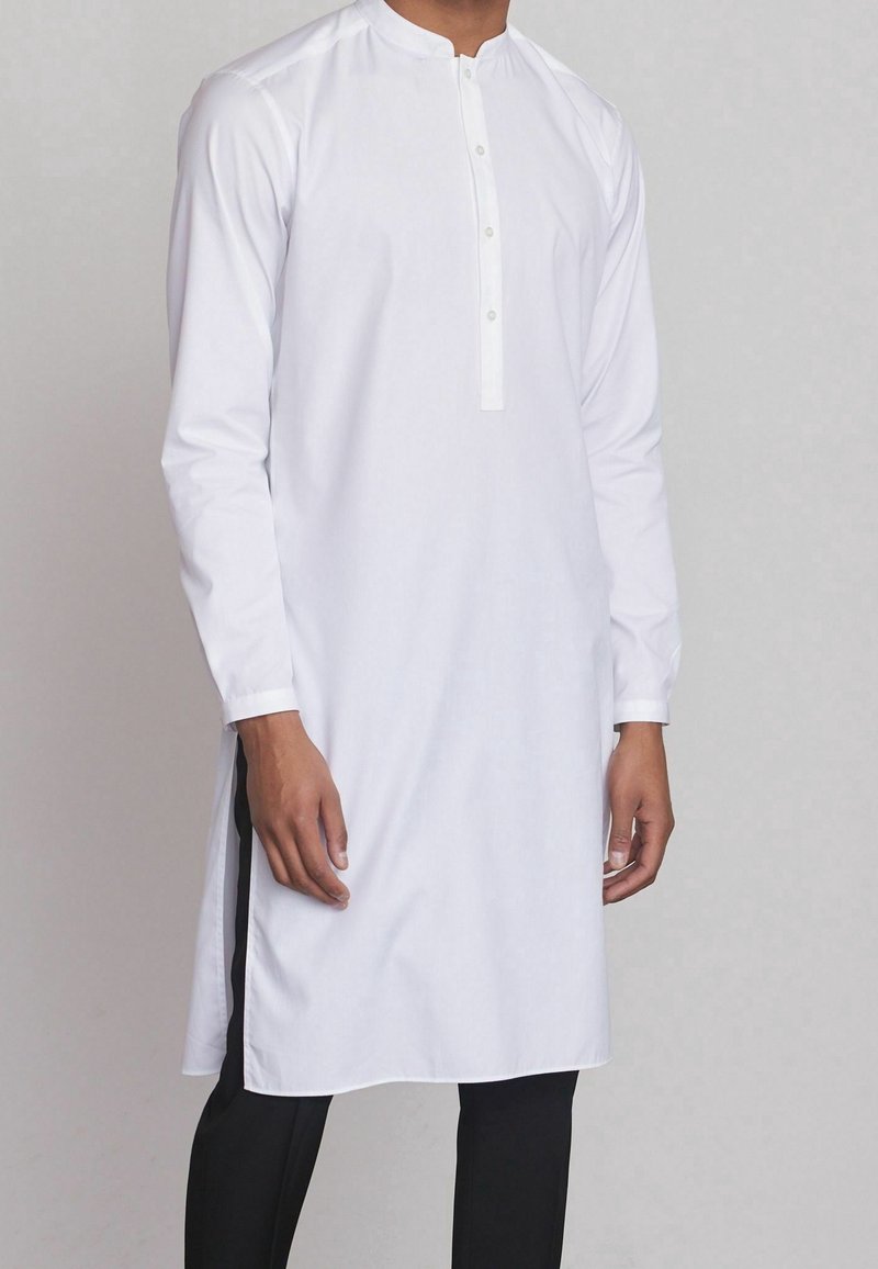 Kurta blanca de algodón con mangas largas, tapeta con botones, cuello redondeado y aberturas laterales; se lleva sobre pantalones negros.