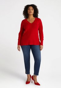 Röd ribbade tröja med scalloped V-hals, matchad med mörkblå slim fit jeans med fransade fållar och guldknapp detaljer. Röda högklackade skor.