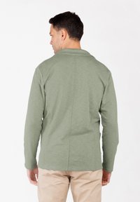 Hombre vestido con una camisa de manga larga verde claro y pantalones beis, de pie con la espalda hacia la cámara contra un fondo blanco liso.