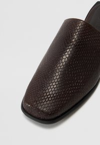 Brown slip-on shoes featuring a textured, hexagonal pattern on the leather, with a rounded toe and a smooth, contrasting black sole.