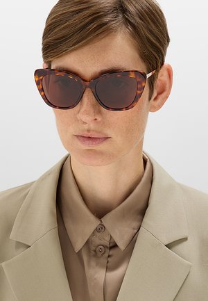 Donna con capelli corti castani che indossa grandi occhiali da sole a gatto tortora e camicia beige con colletto e blazer su sfondo uniforme.