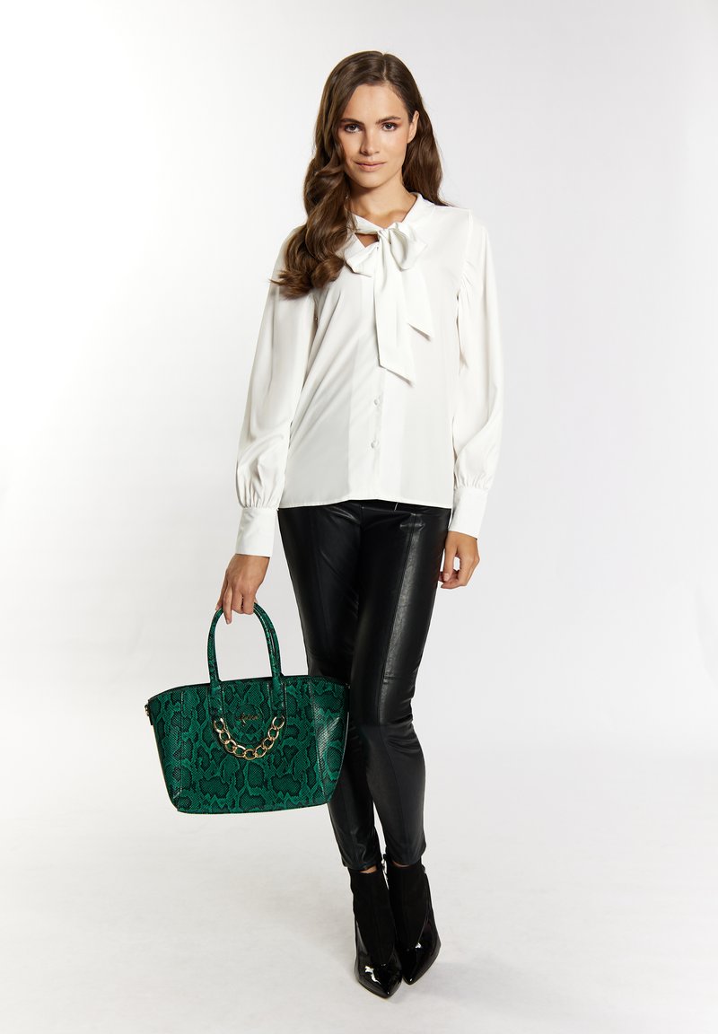 Blusa bianca con dettaglio fiocco, abbinata a pantaloni neri effetto pelle. Tiene una borsa a tracolla verde con motivo serpente e dettagli in catena.