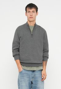 Szara rozpinana bluza z ribbowanymi mankietami i dołem, nałożona na zieloną koszulkę, w połączeniu z jasnoniebieskimi dżinsami. Prosty design, miękka tekstura.