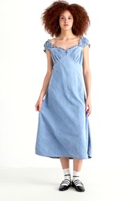 Denim maksi haljina svijetlo plave boje, s rukavima bez ramena i ruffled detaljima, uskim tijelom i lepršavom suknjom, u kombinaciji s crnim cipelama.