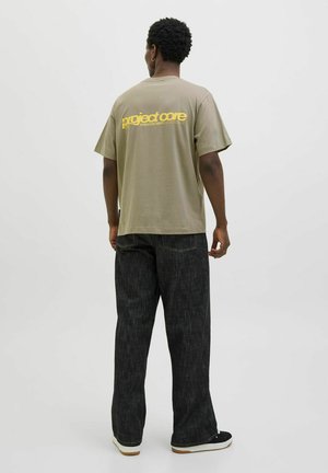Olivengrøn oversized t-shirt med "project core" logo i gul på ryggen, parret med mørkegrå bredbenede jeans og sorte sneakers.