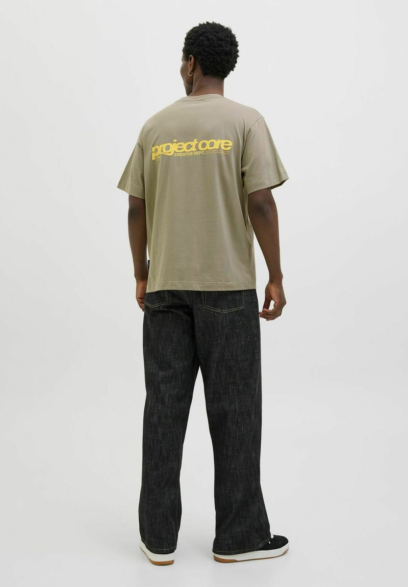Olivengrøn oversized t-shirt med "project core" logo i gul på ryggen, parret med mørkegrå bredbenede jeans og sorte sneakers.