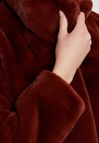 Brązowy płaszcz z futerka syntetycznego z wysokim kołnierzem i szerokimi rękawami. Tekstura jest miękka i puszysta, częściowo leży na dłoni.