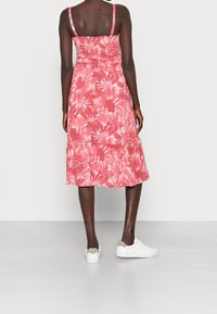 Červené květinové midi šaty s tenkými ramínky, přiléhavým tělem, vrstvami na spodním lemu a lehkým materiálem. Spojené s bílými teniskami na jednobarevném pozadí.