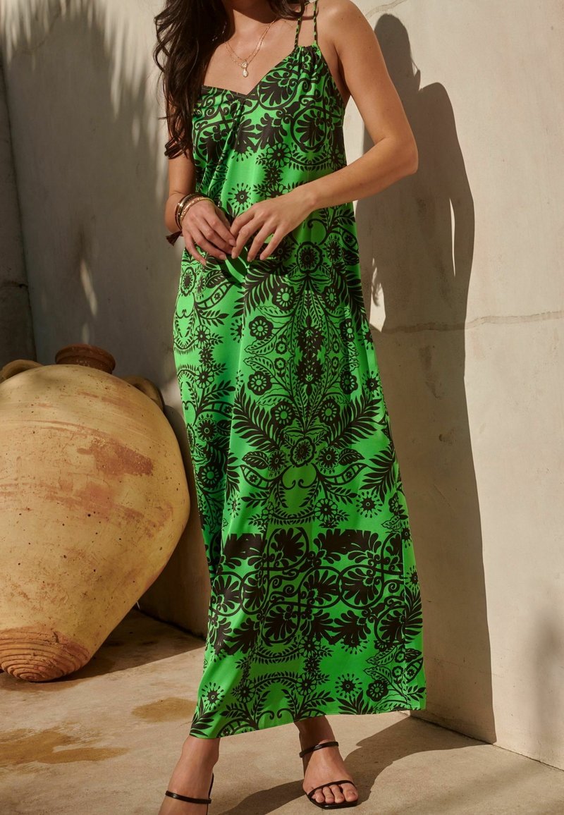 Femme portant une robe maxi sans manches verte avec des motifs floraux noirs, debout à l'intérieur à côté d'un grand pot en céramique, portant des sandales et des bijoux.