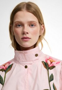 Νεαρή γυναίκα με ανοιχτόχρωμα μαλλιά φοράει ένα ροζ σακάκι με ψηλό γιακά, διακοσμημένο με μεγάλα ροζ και πράσινα λουλουδάτα σχέδια, με το πρόσωπο στραμμένο προς τα εμπρός σε λευκό φόντο.