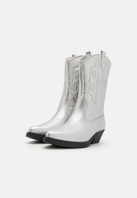Botas de cowboy plateadas hechas de cuero brillante con punteras afiladas, costuras decorativas y tacones bajos negros. Adecuadas para el uso casual.