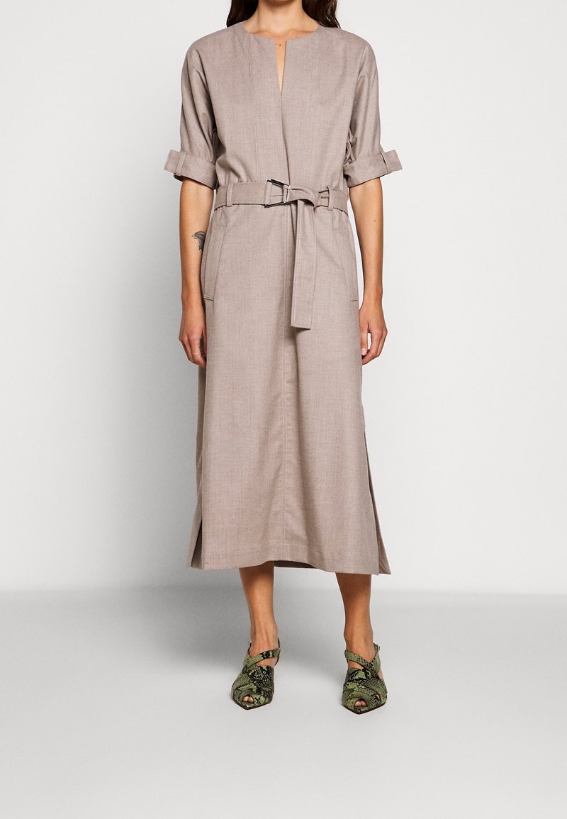 Robe midi en lin beige avec un col en V, des manches retroussées et une ceinture. Présente des fentes latérales et une coupe ample. Associée à des chaussures vertes à motif.