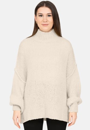 Kobieta z prostymi brązowymi włosami, ubrana w kremowy, oversize'owy sweter z golfem z grubym splotem i czarne spodnie, stojąca na białym tle.