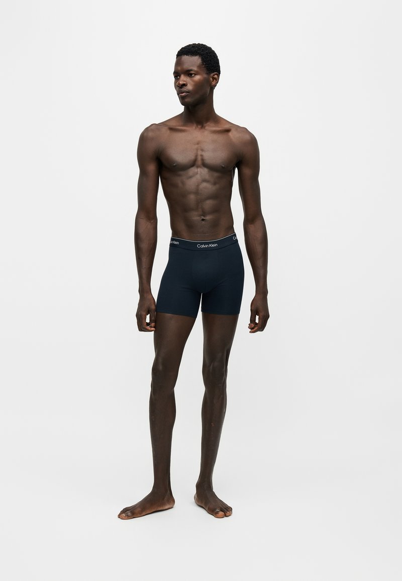 Άνδρας όρθιος ξυπόλητος, φορά σκούρα μπλε boxer Calvin Klein σε απλό φωτεινό φόντο, κοιτάζοντας προς τα αριστερά.