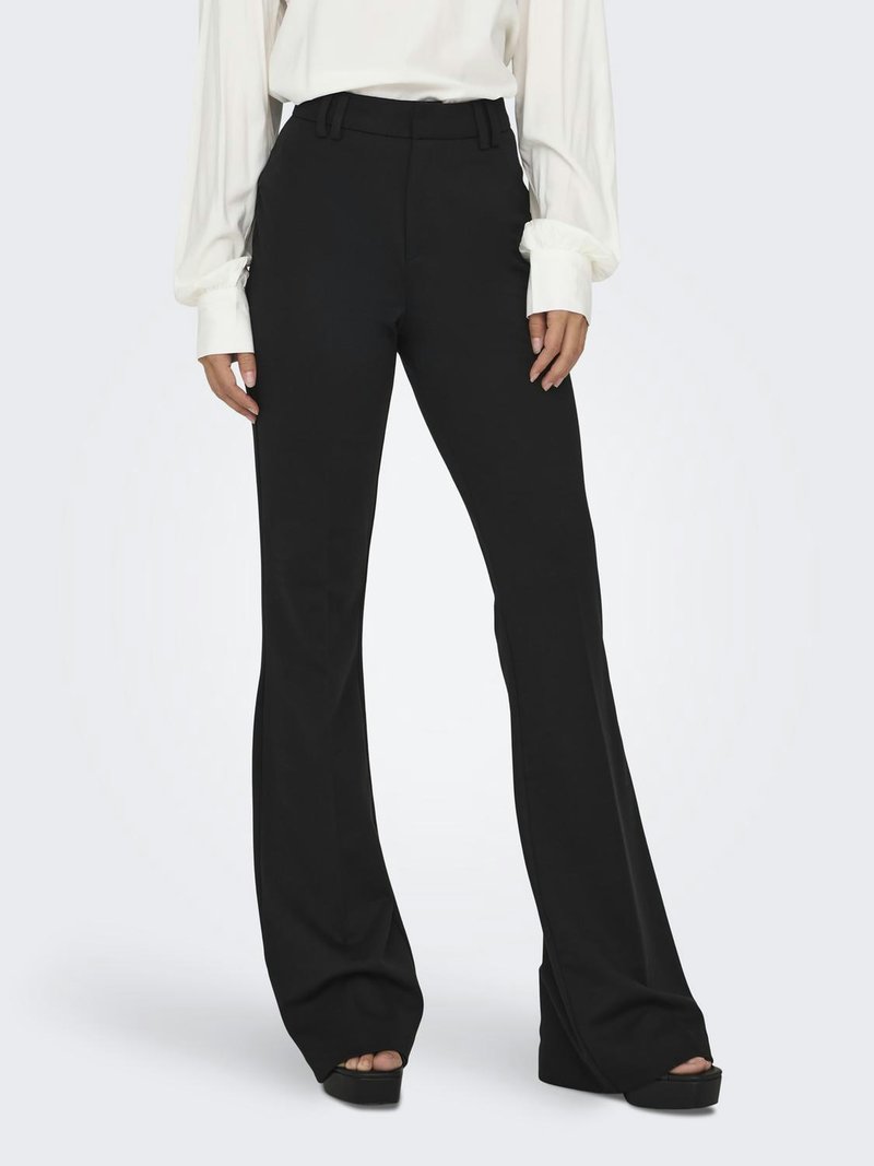 Femme portant un pantalon noir évasé taille haute et un chemisier blanc à manches longues, debout avec les mains détendues le long du corps.