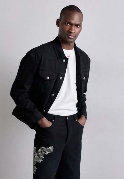 Chaqueta de mezclilla negra con botones metálicos, combinada con una camiseta blanca. Los vaqueros negros presentan un patrón texturizado en la pierna izquierda.