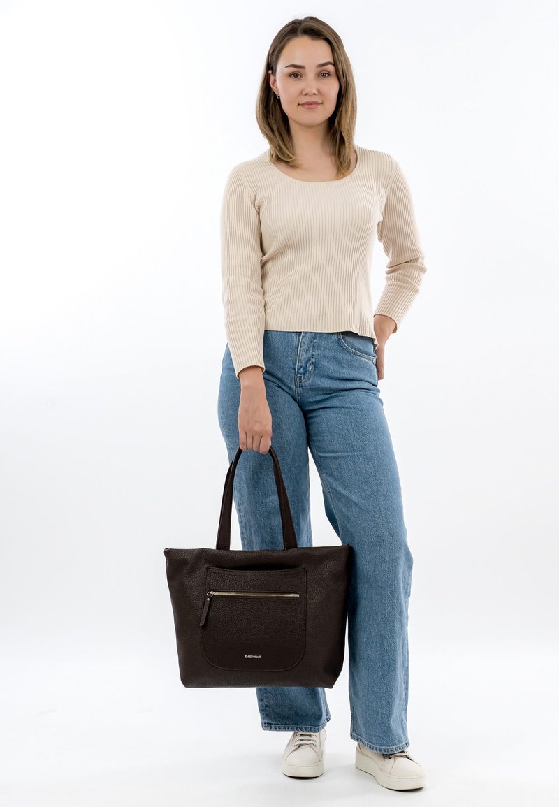 Brūna ādas plecu soma ar teksturētu virsmu, priekšā ar rāvējslēdzēja kabatu un divām stingrām rokturiem. Modelis ģērbies krēmkrāsas džemperī un džinsa biksēs.