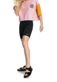 Růžové a oranžové zkrácené tričko s grafickým designem, černé žebrované cyklistické šortky a lesklé černé sandály s nastavitelnými popruhy.