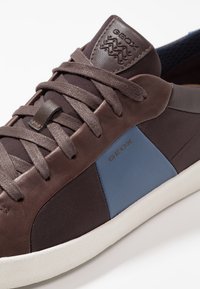 Brązowy sneaker ze skóry i siateczki z niebieskimi akcentami, teksturowanym obszarem na sznurowadła oraz białą gumową podeszwą. Na języku znajduje się wytłoczona logo.