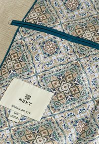Mønstret stof i beige, grøn og blå med intrikate geometriske designs. Inkluderer en hvid etiket med "NEXT" og størrelsesinformation.