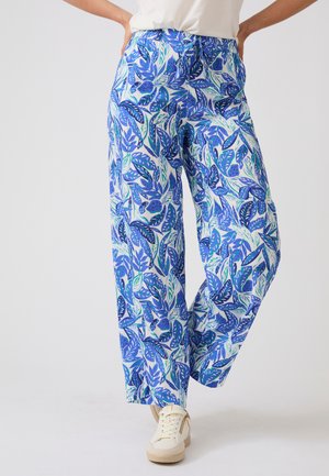 Pantalon à jambes larges avec un motif feuillage bleu et vert, porté avec des baskets blanches et un haut blanc rentré.