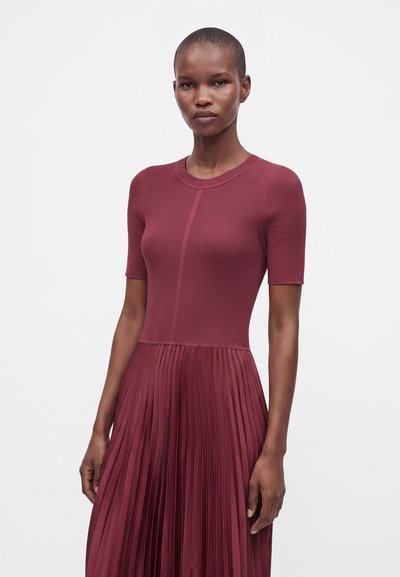 Femme aux cheveux courts portant un haut bordeaux moulant à manches courtes côtelées et une jupe plissée assortie sur un fond clair uni.