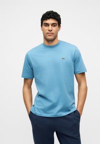 Άνδρας με σγουρά μαλλιά που φοράει ανοιχτό μπλε κοντομάνικο πουκάμισο με μικρό πράσινο λογότυπο, στέκεται με τα χέρια στις τσέπες μπλε σκούρου παντελονιού.