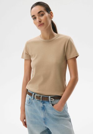 Kobieta z ciemnymi włosami związanymi do tyłu, ubrana w beżowy t-shirt włożony w jasnoniebieskie jeansy z brązowym paskiem, stoi z jedną ręką w kieszeni.