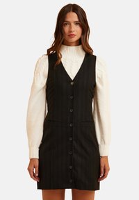 Abito nero a righe con un design aderente, scollo a V, chiusura frontale con bottoni e due tasche, indossato sopra un maglione a collo alto a coste color crema.