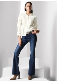 Witte blouse met lange mouwen en kantdetails, gecombineerd met donkerblauwe flare jeans en beige enkellaarsjes met een hak op witte treden.