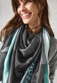 Gran bufanda gris con acentos turquesa, que presenta un patrón texturizado y bordes con flecos, drapeada alrededor de un cuello. Material suave.