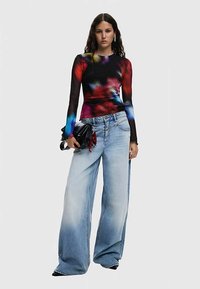 Langärmliges, tailliertes Oberteil mit einem mehrfarbigen, abstrakten Muster. Locker sitzende, weitgeschnittene Jeans in Hellblau mit einem ausgewaschenen Effekt. Schwarze Handtasche als Akzent.