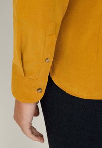 Camisa de pana amarilla con dos puños abotonados. La textura de la tela es acanalada, creando una apariencia suave. Tiene un dobladillo curvado notable y un cuello redondeado.
