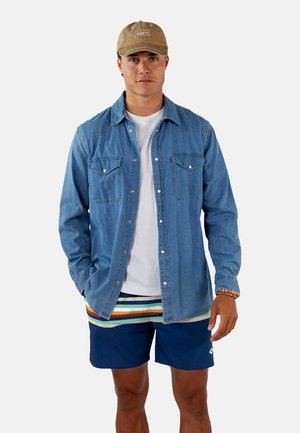 Denim shirt in blue with two chest pockets, white t-shirt underneath, multicolored striped shorts, and a beige cap with text.