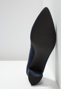 Escarpin bleu marine en daim avec un bout pointu, doté d'une semelle en caoutchouc noir avec des rainures horizontales pour une meilleure adhérence.