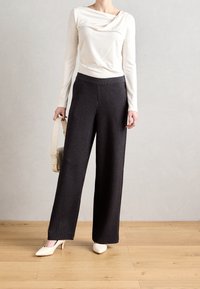 Witte lange mouwen top met gedrapeerde halslijn, gecombineerd met zwarte pinstriped wijde broek en crèmekleurige enkellaarsjes met puntige neus, met een lichte handtas in de hand.