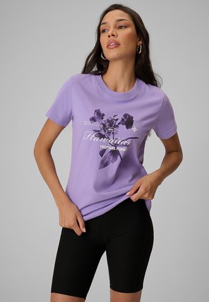 Kobieta w fioletowej koszulce z kwiatowym nadrukiem i napisem "Hawaiian Tropical Flower", ubrana w czarne prążkowane szorty, stoi na jednolitym tle.