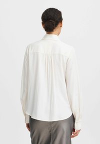 Blouse à manches longues de couleur crème clair avec une texture douce. Elle possède un col, des poignets boutonnés et un détail froncé à l'arrière pour un ajustement décontracté.