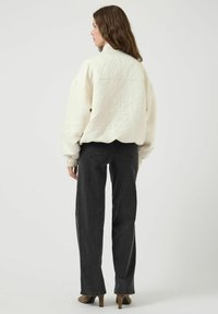 Giacca crema trapuntata con colletto alto, polsini elastici, abbinata a jeans scuri a vita alta e tacchi taupe, vista da dietro.