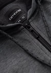 Sudadera con cremallera negra y gris con un patrón texturizado, que cuenta con capucha ajustable con cordón, cremallera frontal y una etiqueta de marca visible en el interior.