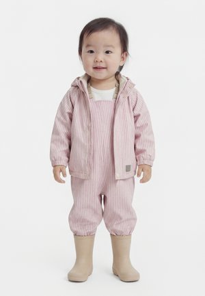 Νήπιο που φοράει ροζ ριγέ μπουφάν και ταιριαστά παντελόνια με μπεζ αδιάβροχες μπότες, στέκεται και κοιτάει μπροστά, με ελαφρύ χαμόγελο.