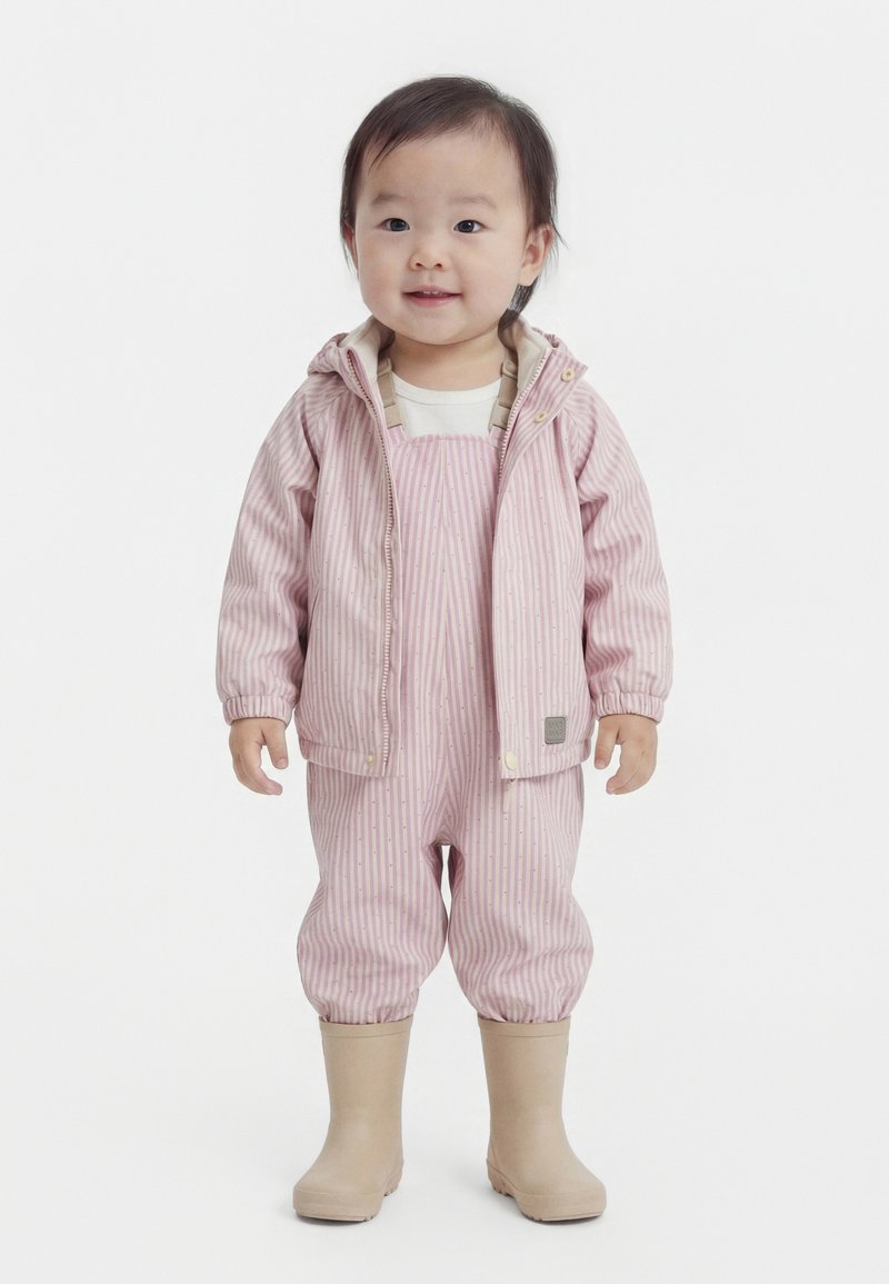 Νήπιο που φοράει ροζ ριγέ μπουφάν και ταιριαστά παντελόνια με μπεζ αδιάβροχες μπότες, στέκεται και κοιτάει μπροστά, με ελαφρύ χαμόγελο.