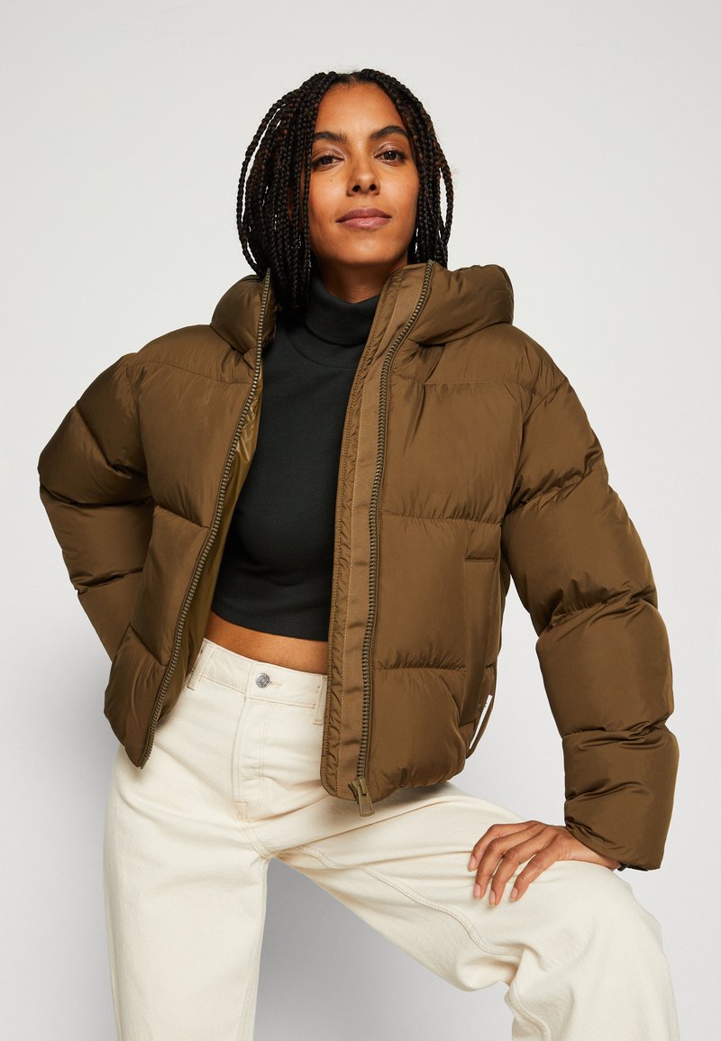 Braune Pufferjacke mit Kapuze, mit einem gesteppten Design und einem Frontreißverschluss. Kombiniert mit einem schwarzen Rollkragenpullover und cremefarbenen Hosen.